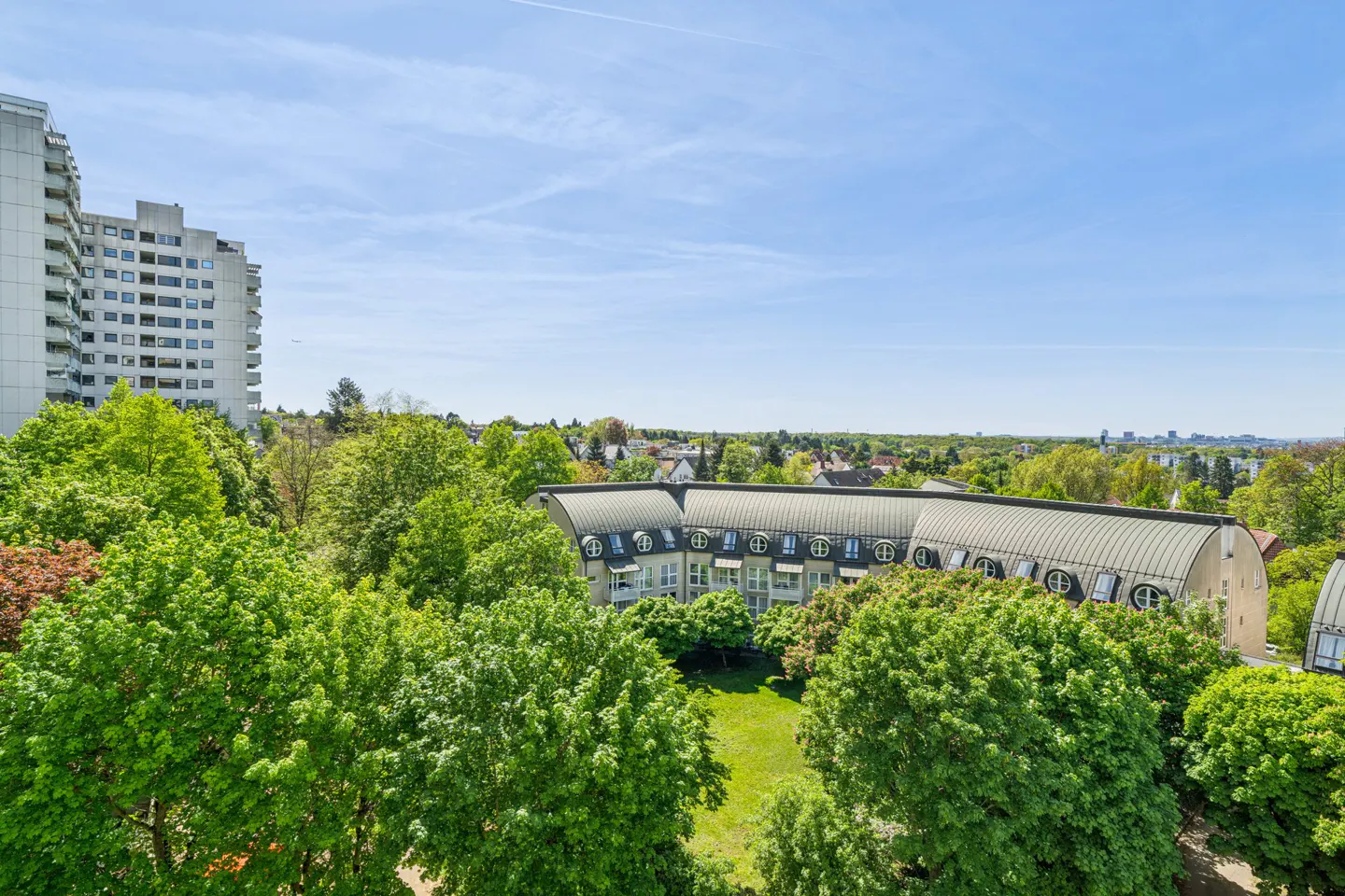 View of a city with green trees, a tall apartment building, and a curved roof building under a blue sky.