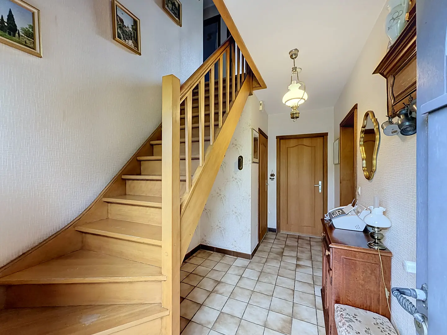 Entryway with a wooden staircase, tiled floor, a wooden cabinet with a lamp, and a closed wooden door in the background.