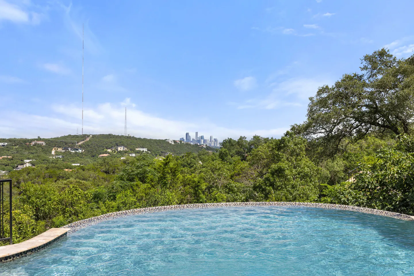Infinity pool overlooking a lush green hillside with a distant city skyline under a bright blue sky.