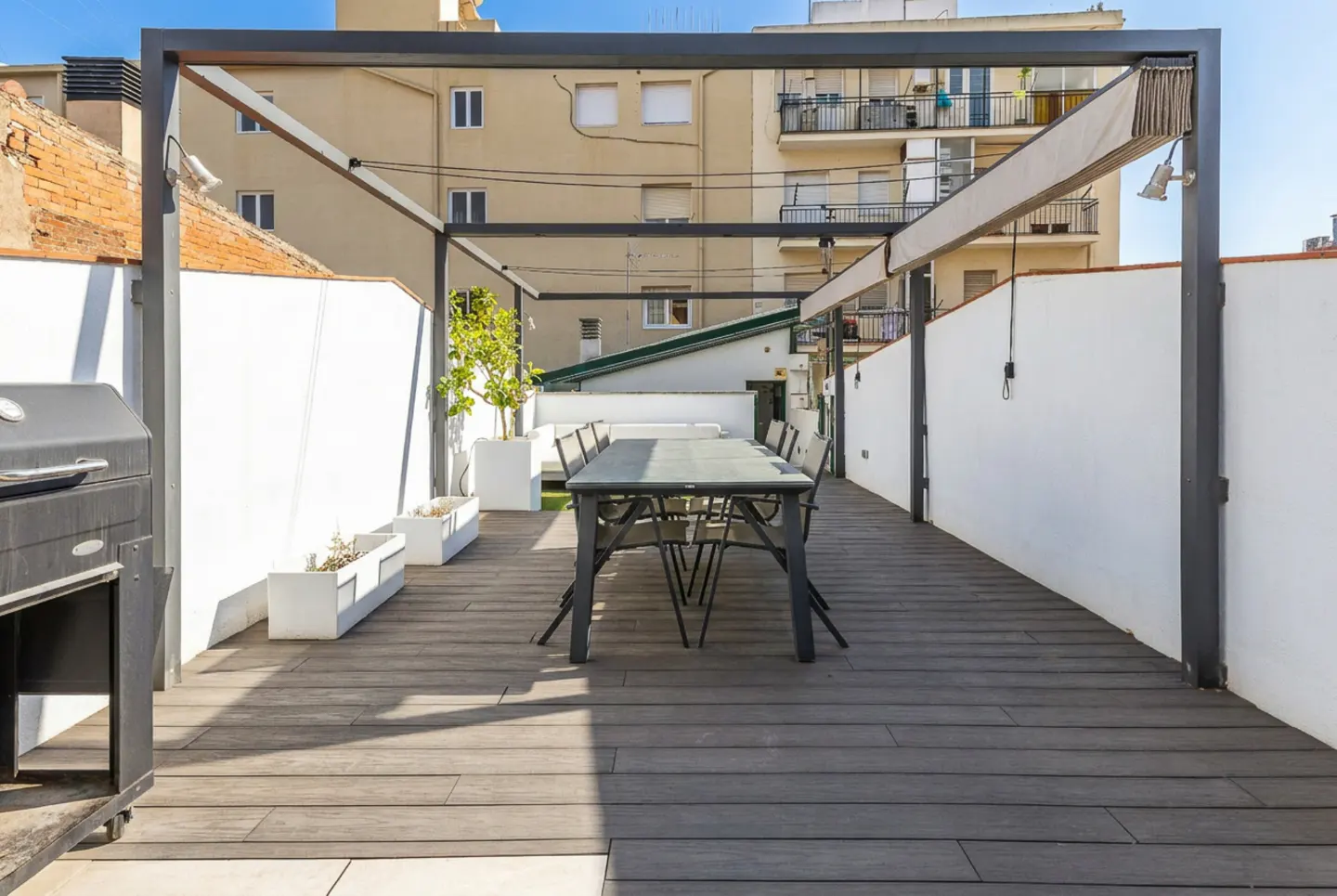 Outdoor patio with gray wood flooring, a black metal pergola, a dining table, and a grill. White walls and planters add contrast.