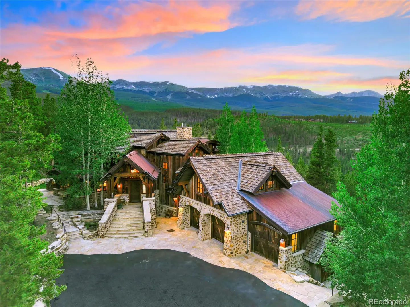 A rustic mountain home with stone accents, a wood roof, and a three-car garage, set against a backdrop of mountains and a colorful sunset.
