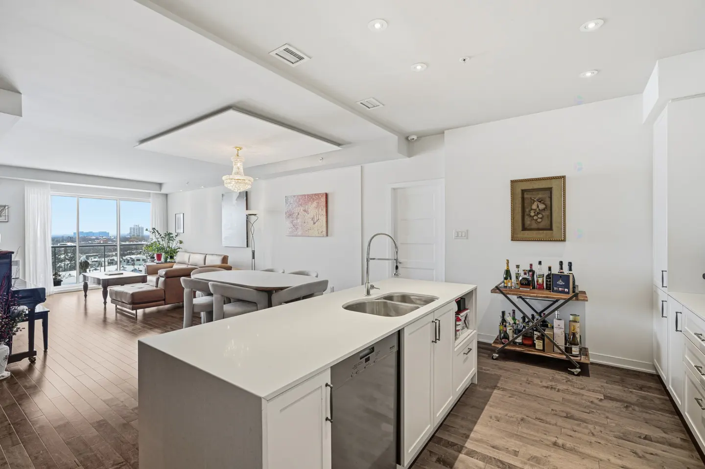 Bright, open-concept condo with white walls, wood floors, and a kitchen island. Living area with brown leather sofa and city view.