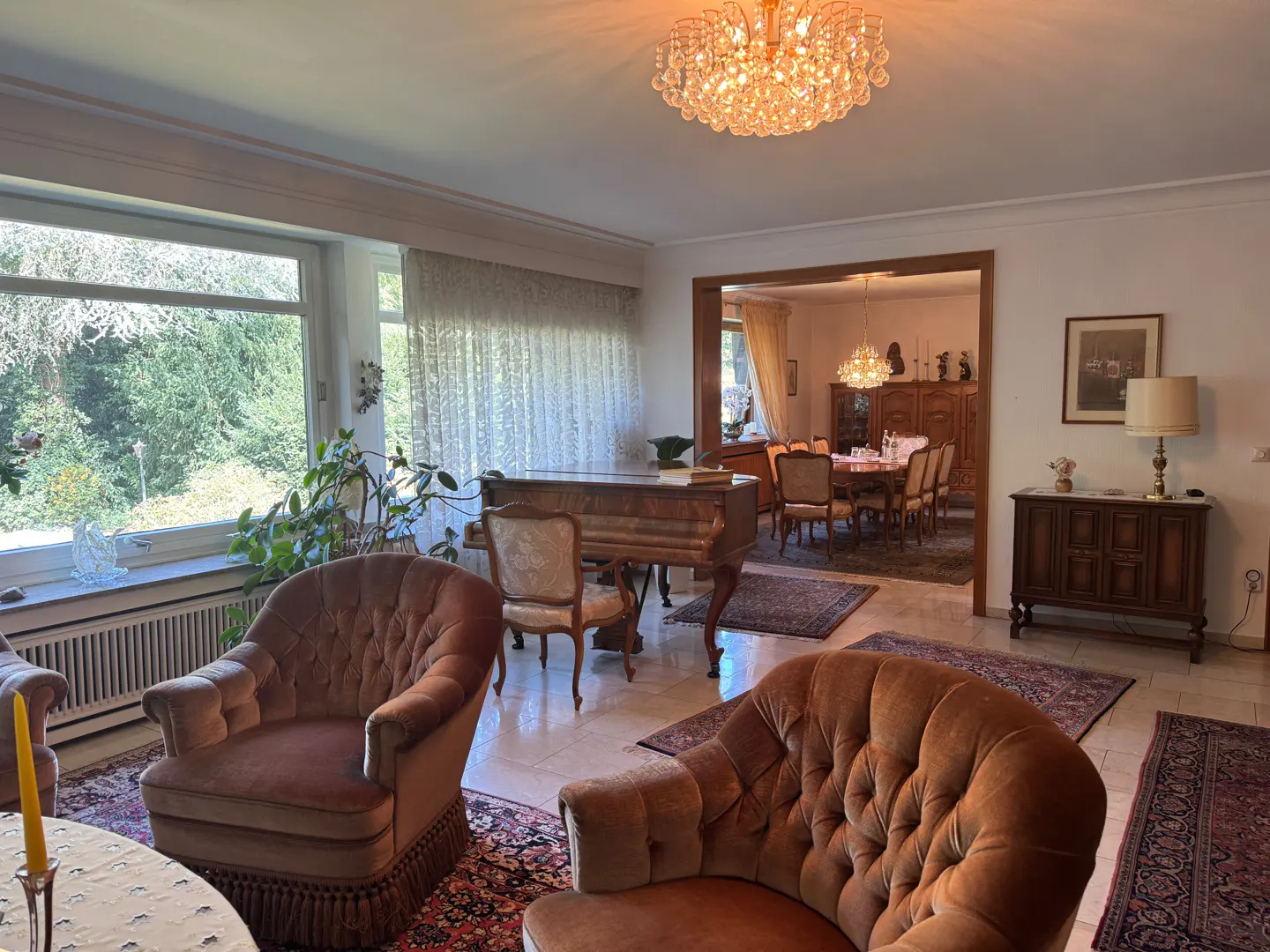 A living room with brown armchairs, a piano, and a dining room in the background. A chandelier hangs from the ceiling.