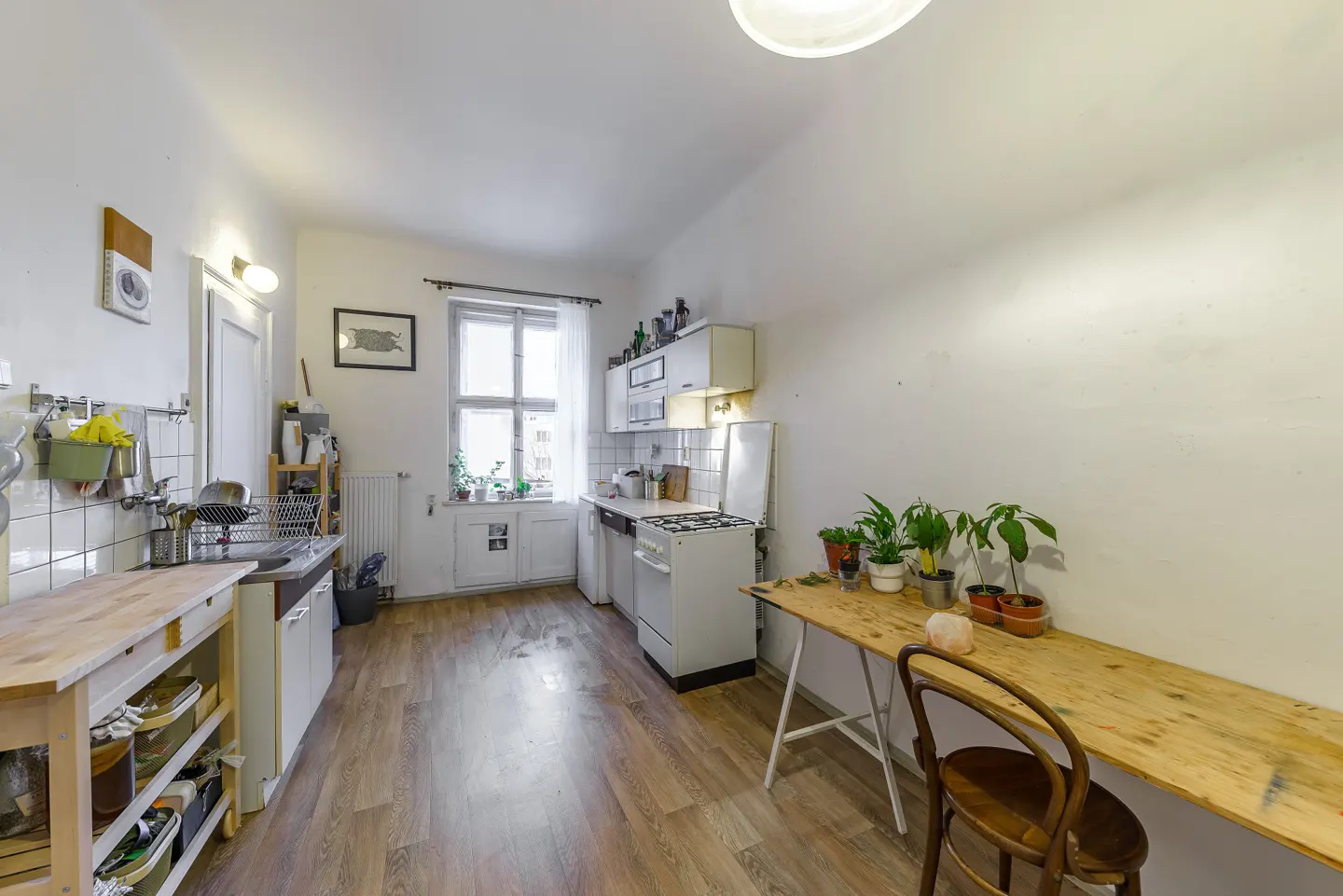 Bright kitchen with white walls, wood floors, and a window. A wooden table with plants sits against one wall.