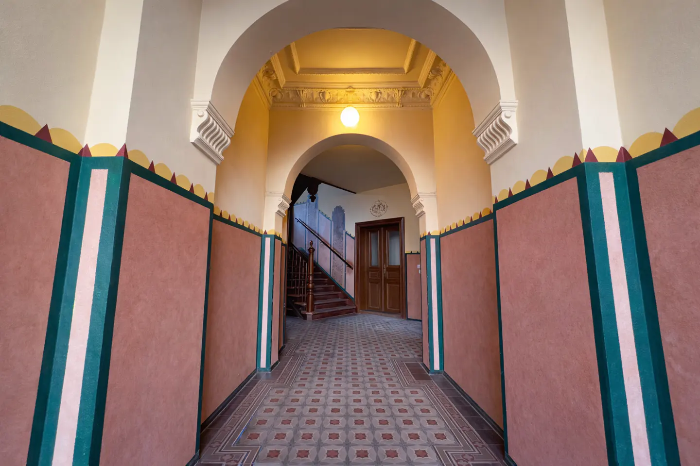 A hallway with arched ceilings, patterned tile floors, and walls painted in pink, green, and yellow with decorative trim.