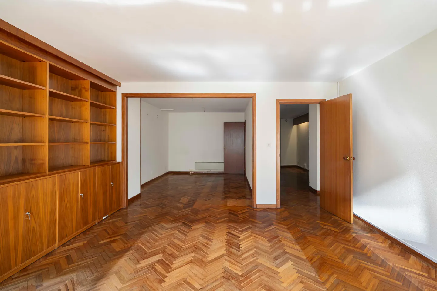 Bright room with herringbone wood floor, white walls, and a large wooden bookcase. Open doorway leads to another room.