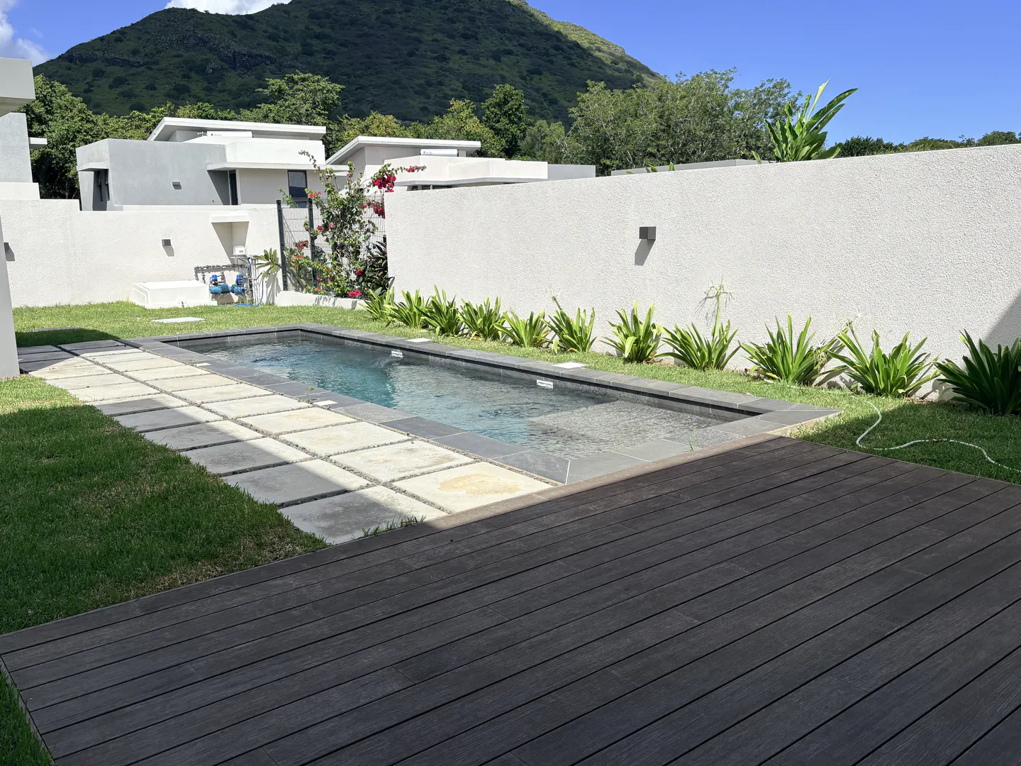 A rectangular swimming pool with a wooden deck and stone pavers, surrounded by a white wall and green lawn. A mountain is in the background.