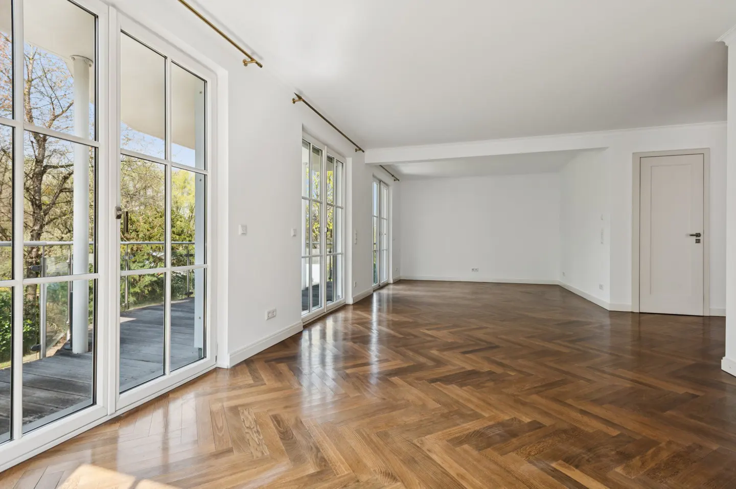 Bright, empty room with herringbone wood floors, white walls, and large windows overlooking greenery.