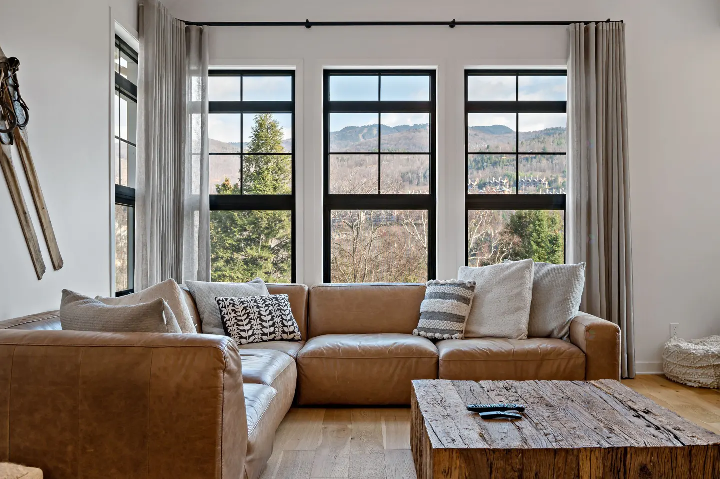 Living room with a brown leather sectional sofa, rustic wood coffee table, and mountain view through black-framed windows.