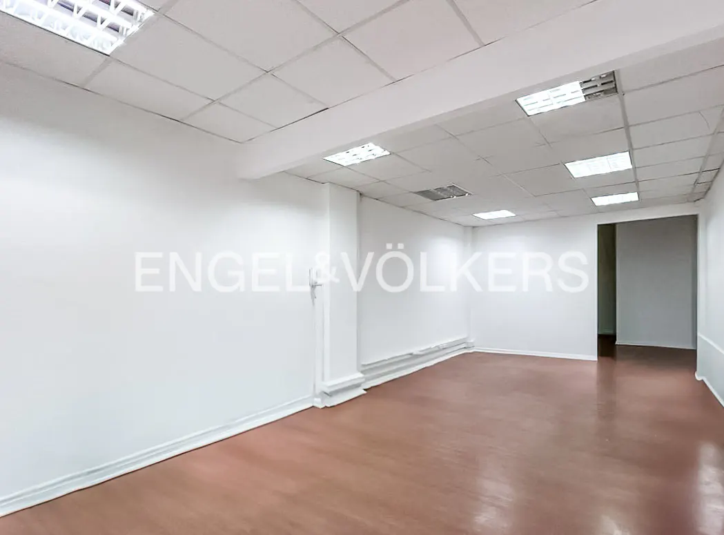 Empty commercial space with white walls, brown floor, and a white grid ceiling with fluorescent lights. Engel & Völkers logo on the wall.