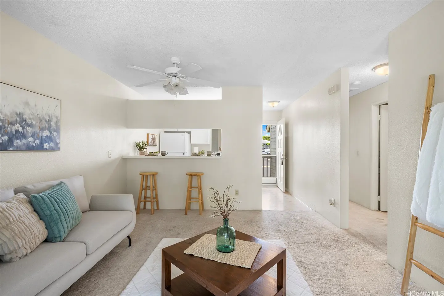 Living room with a light gray sofa, teal pillow, wooden coffee table, and a breakfast bar with two stools.