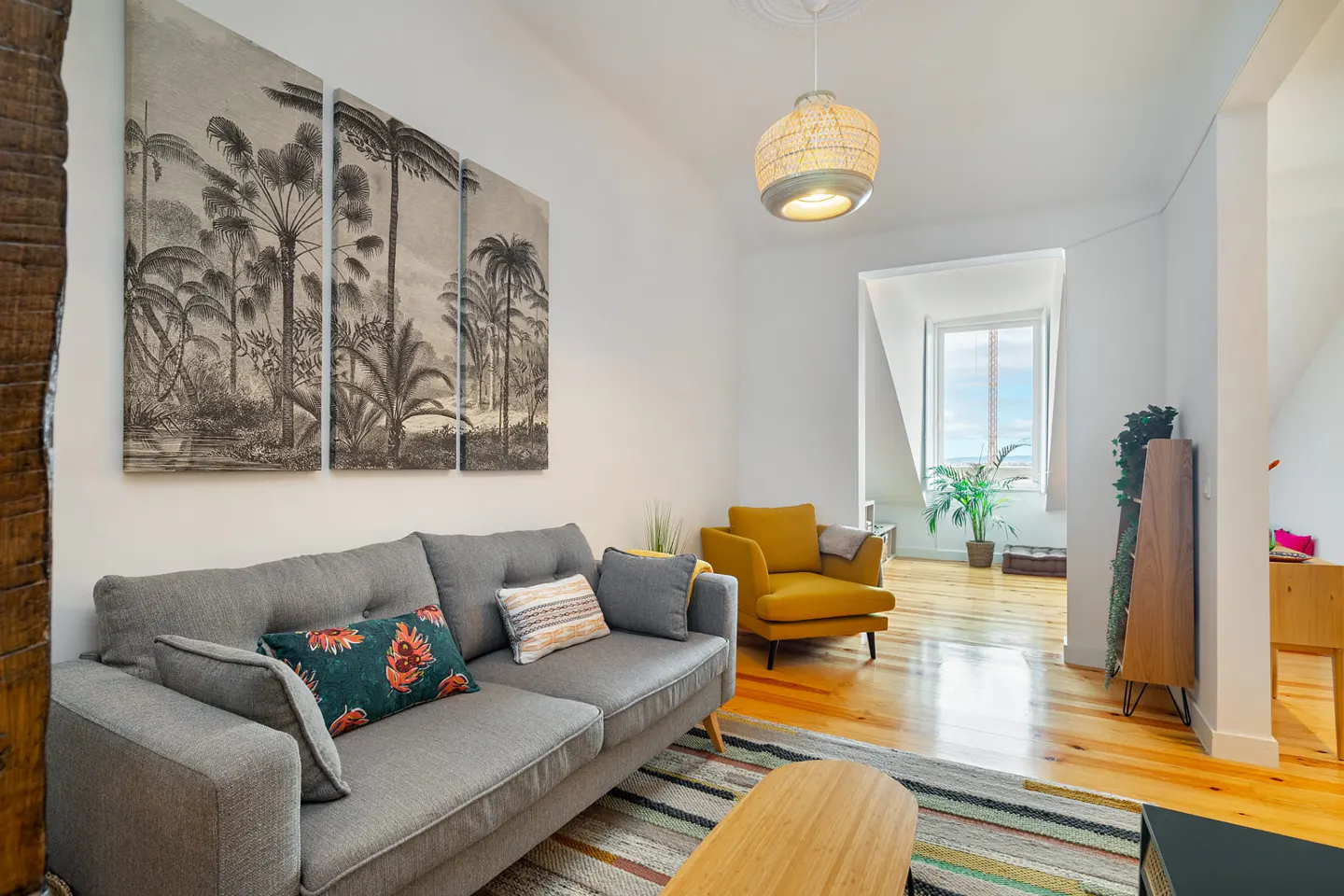 Bright living room with gray sofa, yellow chair, and tropical artwork. Hardwood floors and a window with a view.
