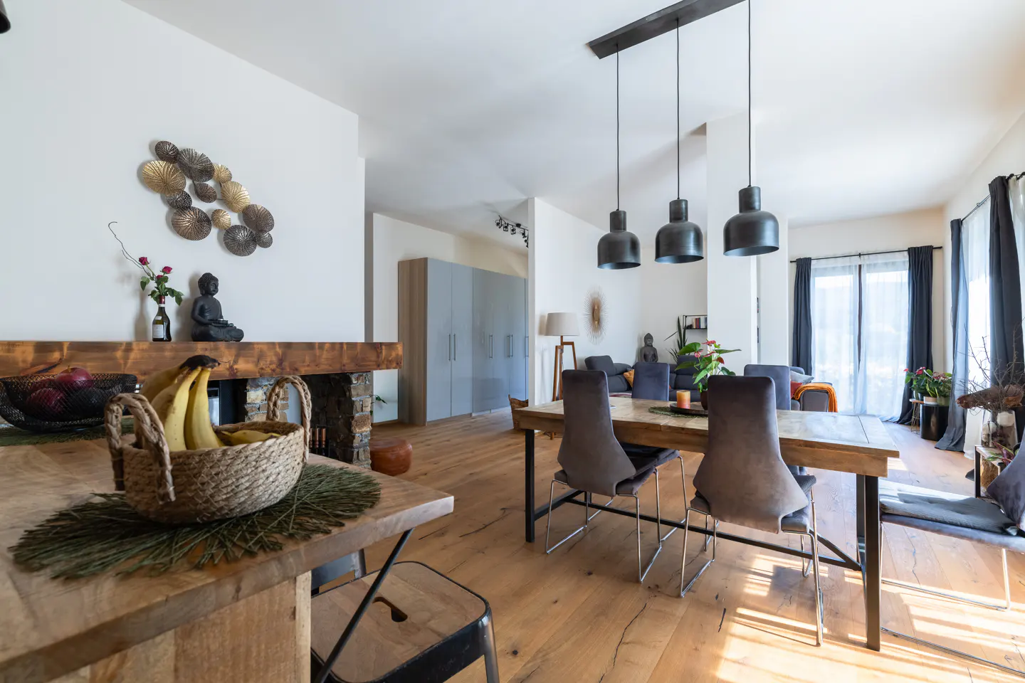 A bright, modern dining room with a wooden table, gray chairs, and black pendant lights. A fireplace with a wooden mantel is visible in the background.