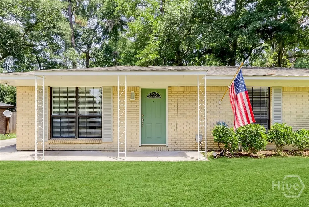 Tan brick ranch-style house with a green door, white porch columns, and an American flag waving in front.