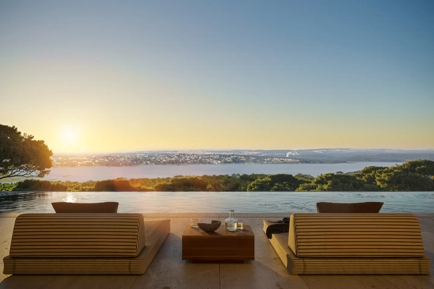 Two lounge chairs face an infinity pool overlooking a bay at sunset. A small table sits between them with a glass bottle and bowl.