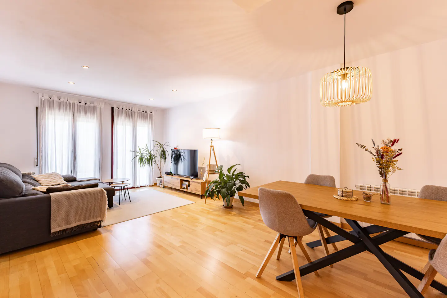 Bright living room with wood floors, a gray sofa, a wooden dining table with gray chairs, and large windows.