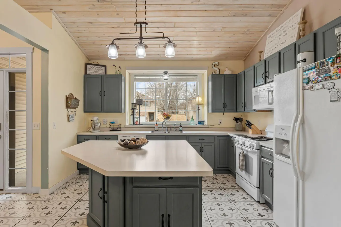 A kitchen with gray cabinets, a white island, and a white refrigerator. The floor has a patterned tile design. A window overlooks a yard with trees.