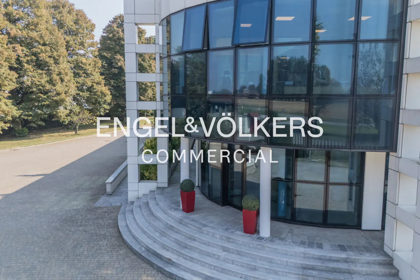Modern office building with a curved glass facade, stone steps, and red planters, branded with Engel & Völkers Commercial.