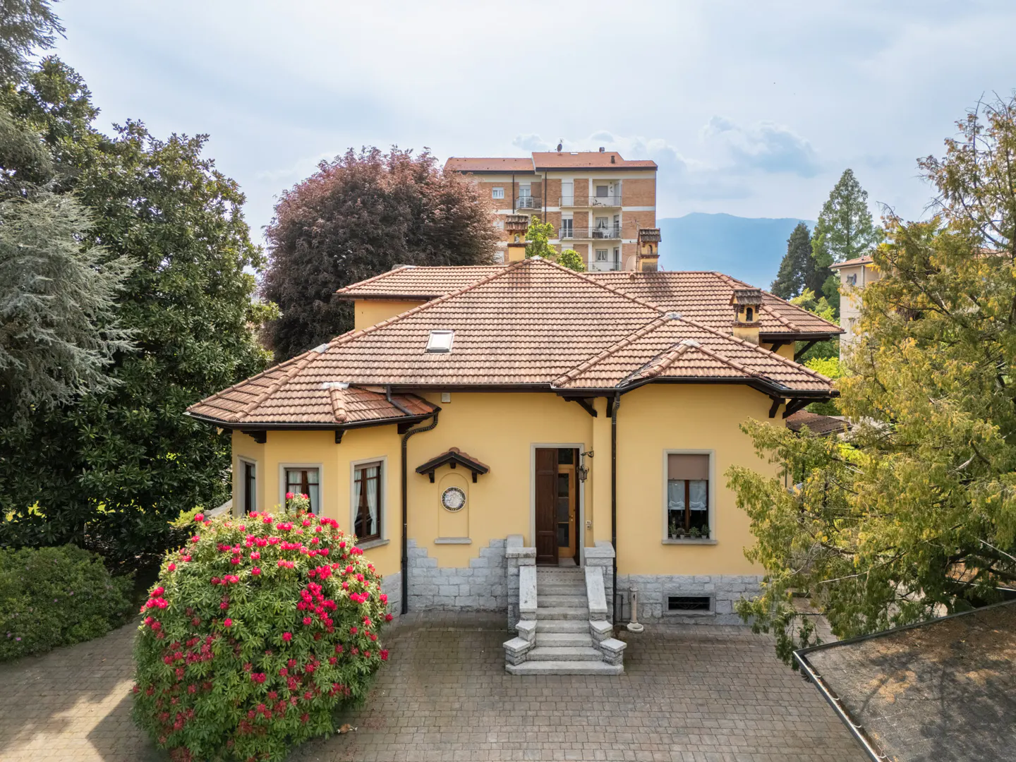A yellow two-story house with a brown tile roof, surrounded by trees and a red flowering bush.