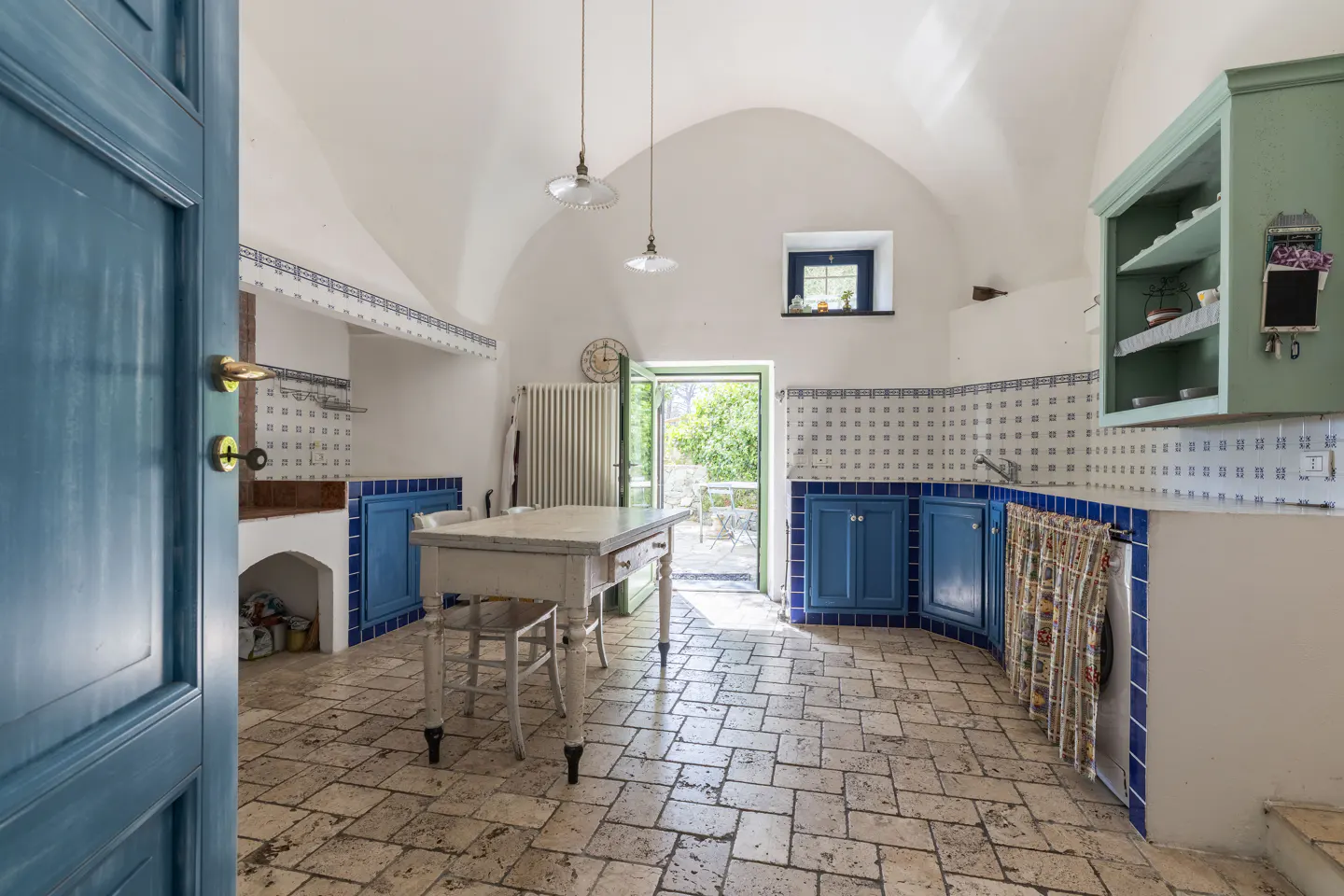 A bright kitchen with blue cabinets, tiled walls, and a stone floor. A white table sits in the center, and a door leads to an outdoor area.