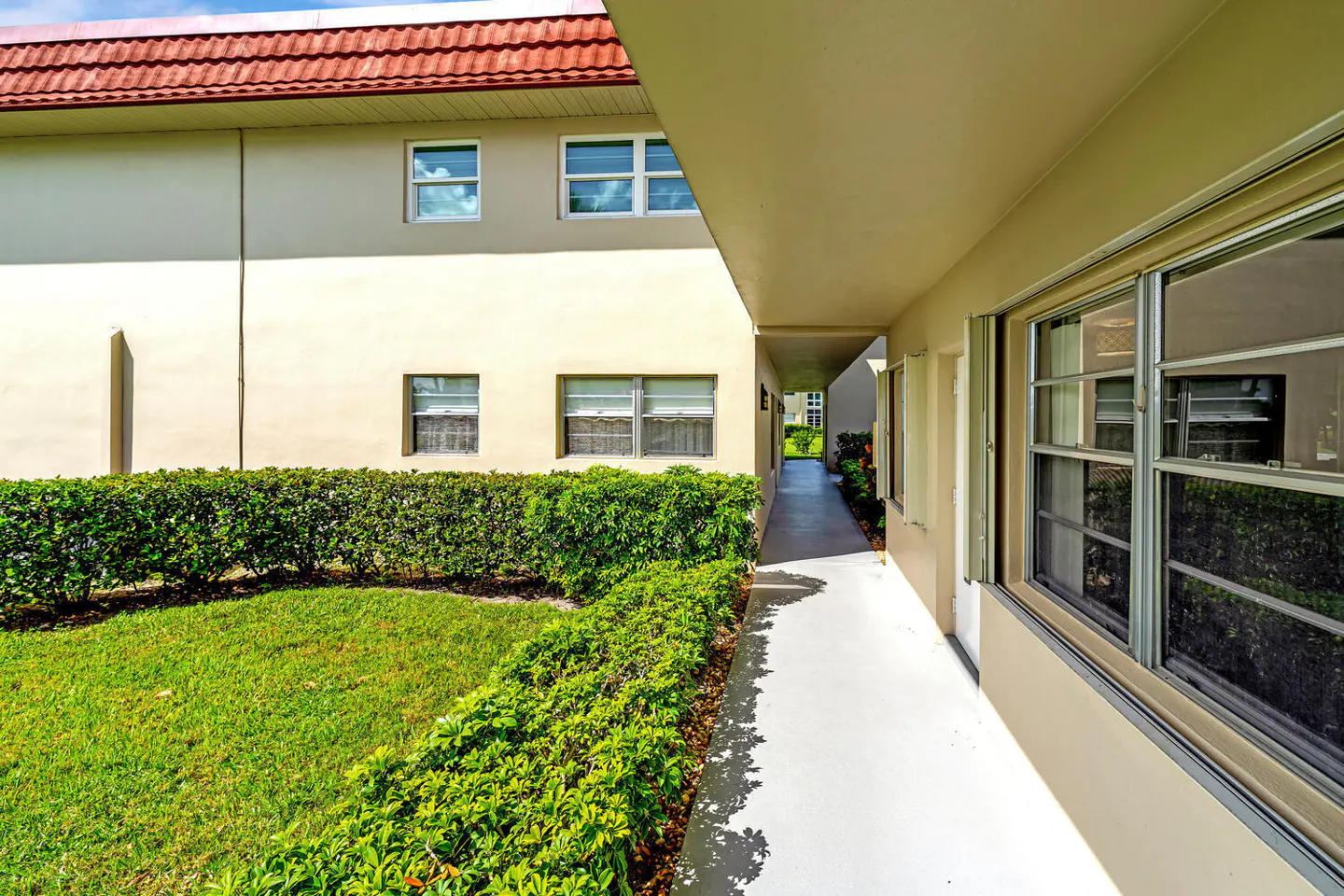 Exterior view of a beige apartment building with a red tile roof. A walkway is lined with green bushes and windows.
