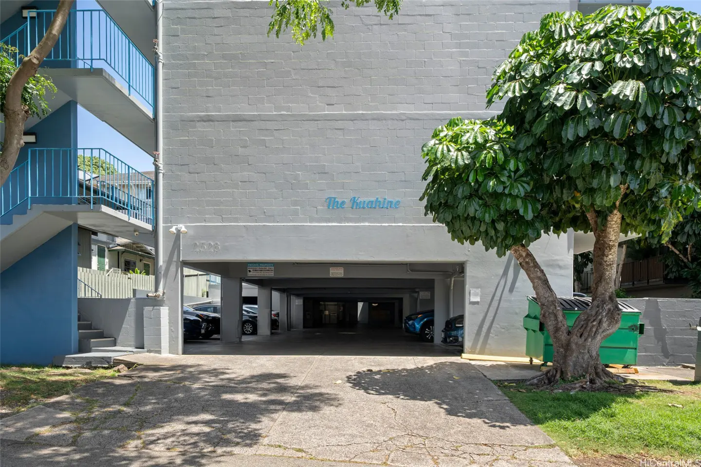 Exterior view of "The Kuahine" building with parking garage, stairs, and a tree. The building is painted gray.