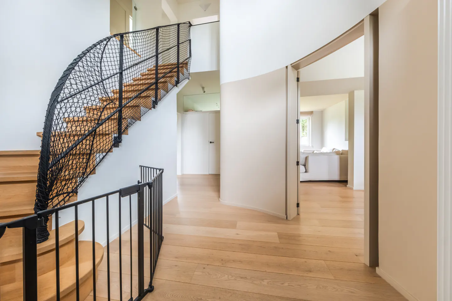 Interior view of a modern home featuring a wooden staircase with a black net railing, light wood floors, and an open doorway leading to a living room.