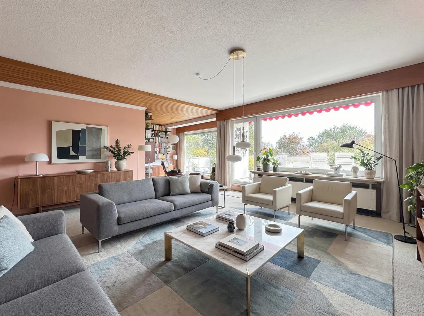 A bright living room with gray sofas, white armchairs, and a marble coffee table on a patterned rug. Large windows offer a view of greenery.