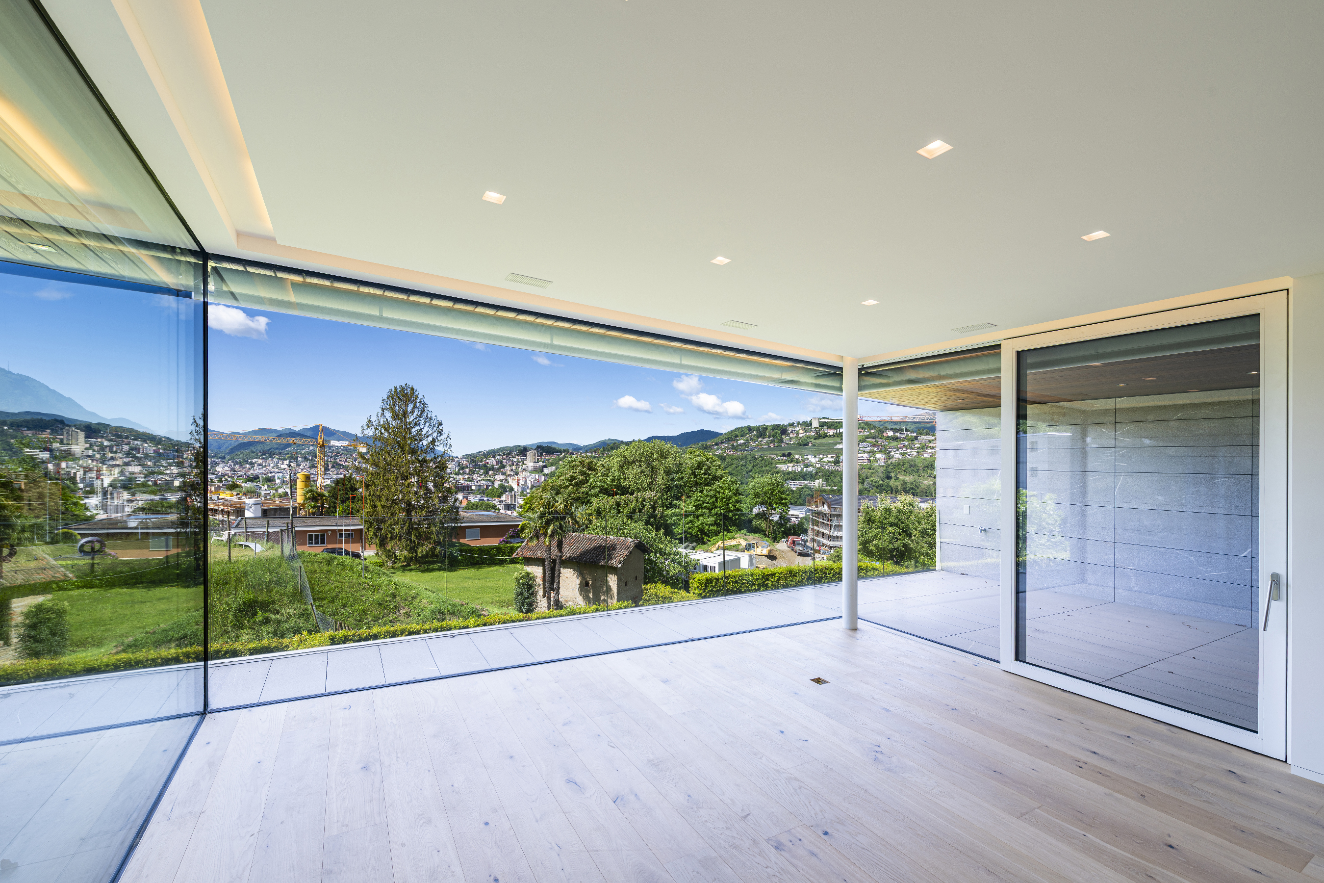 Modern room with light wood floors and floor-to-ceiling glass walls overlooking a lush valley, trees, and distant mountains.