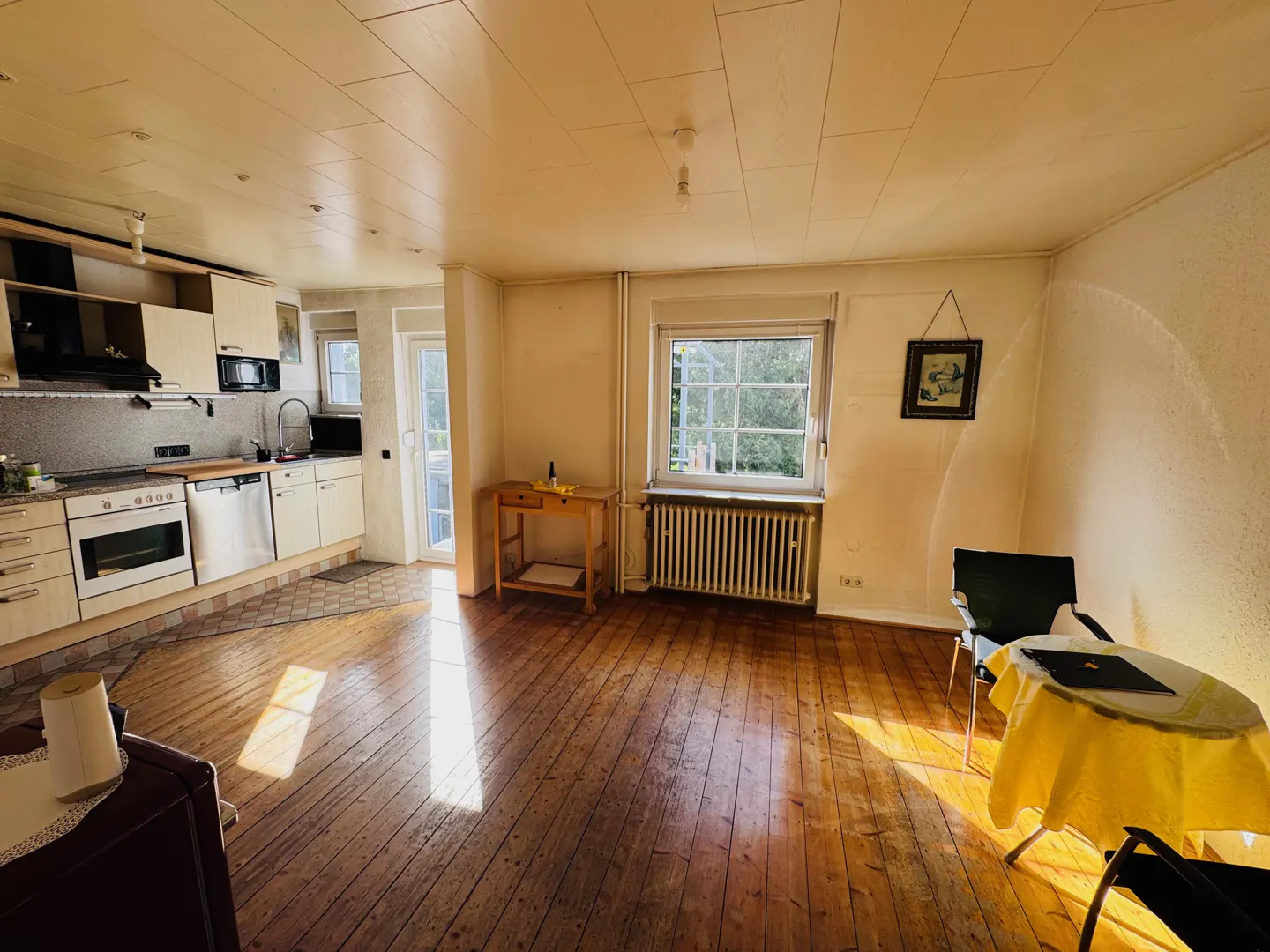Sunlit apartment interior with wood floors, a kitchen area, and a table with a yellow cloth. A window overlooks greenery.