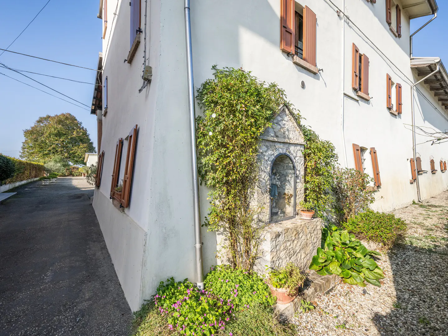 Exterior view of a white building with brown shutters and a stone shrine with plants.