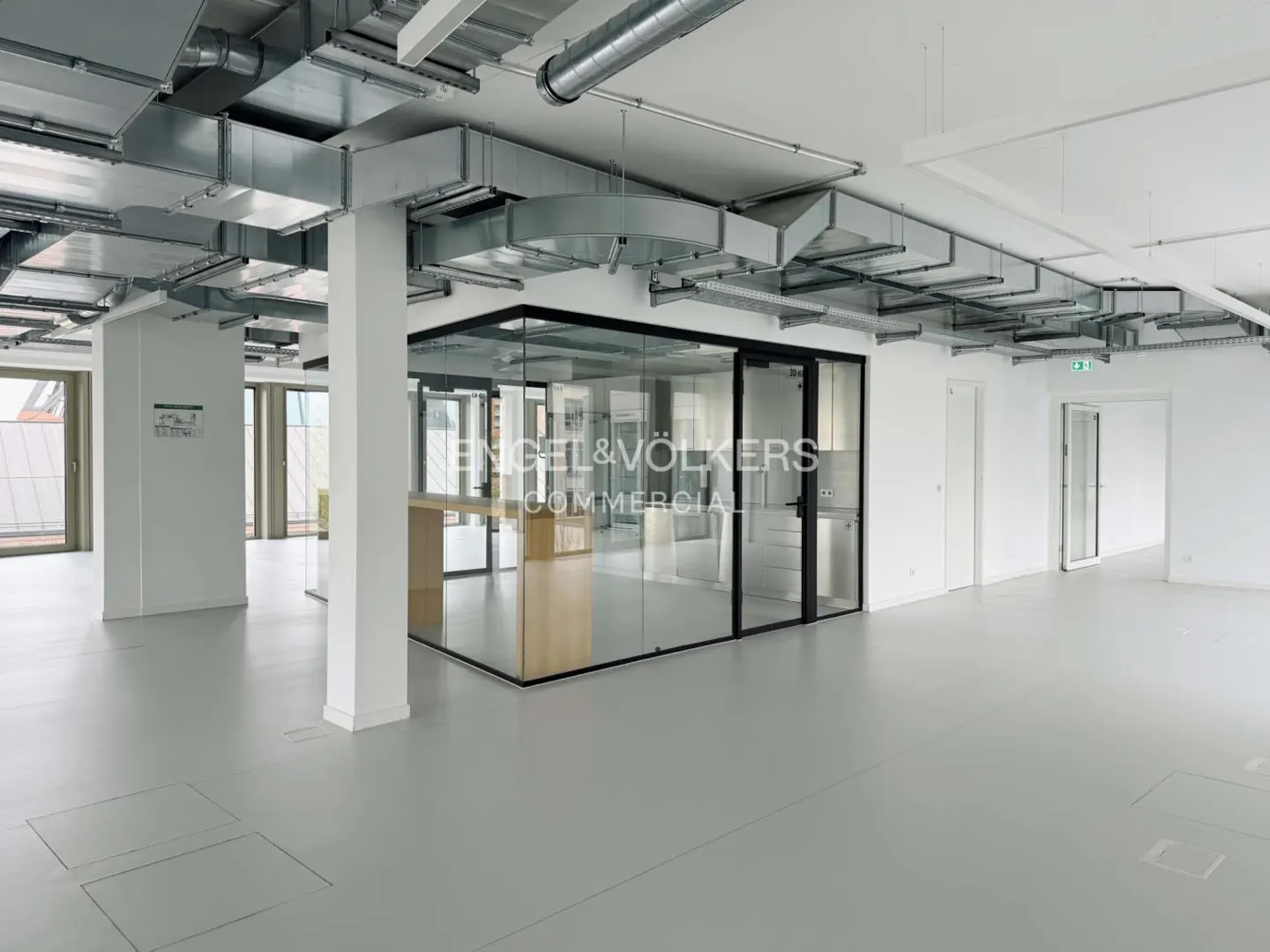 Empty office space with gray floors, white walls, and exposed ductwork. A glass-walled office with the Engel & Völkers logo is in the center.