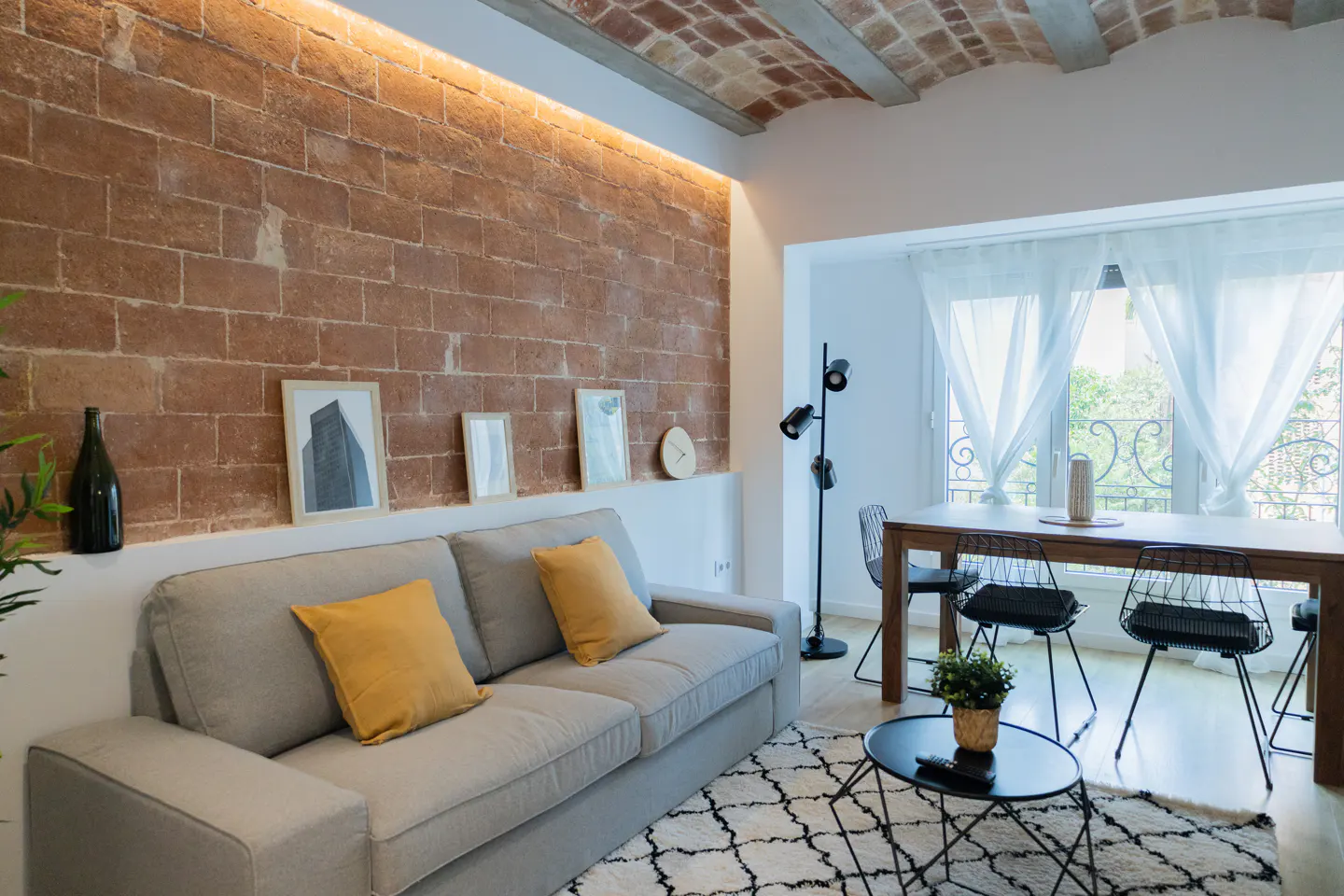 Modern living room with exposed brick wall, vaulted ceiling, grey sofa with yellow pillows, and a dining area by a window.
