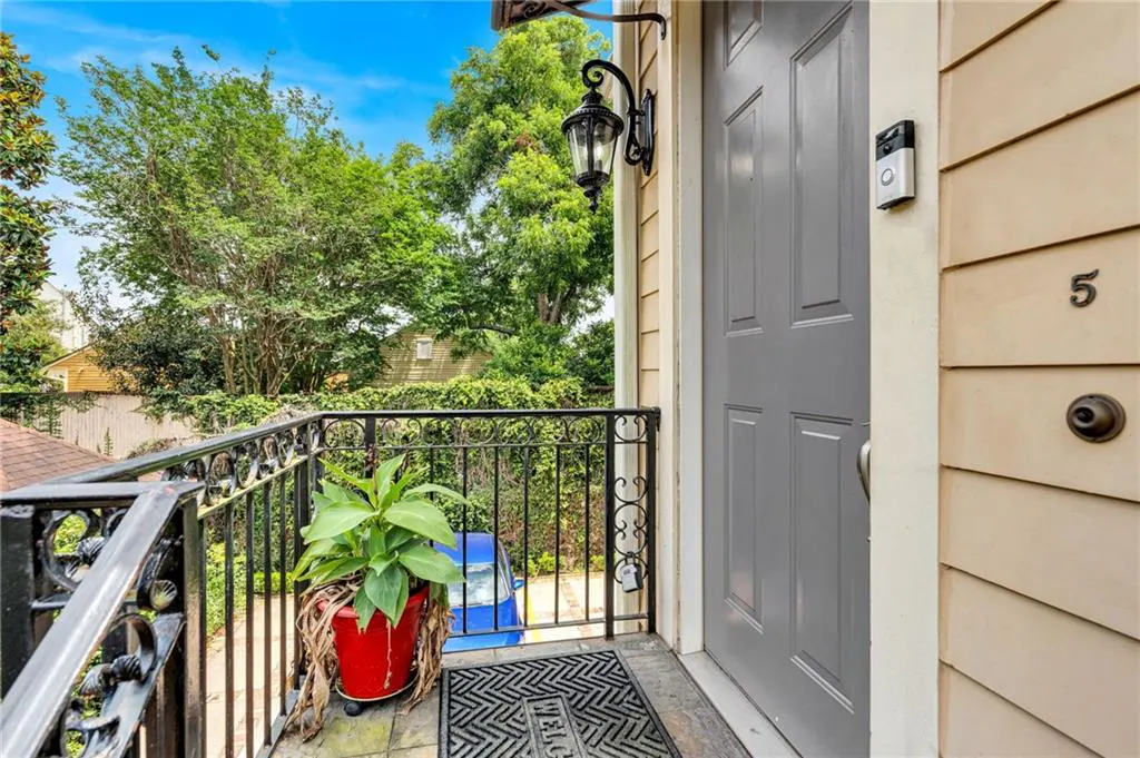 A gray front door with a doorbell camera, a potted plant, and a black metal railing overlooking a tree-lined street.