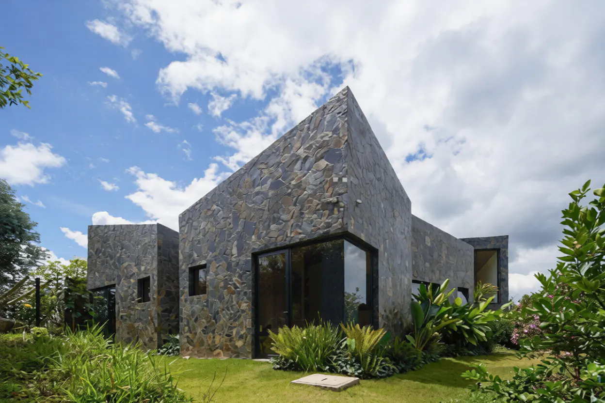 Modern home with dark stone exterior, large glass windows, and lush green landscaping under a blue sky with clouds.