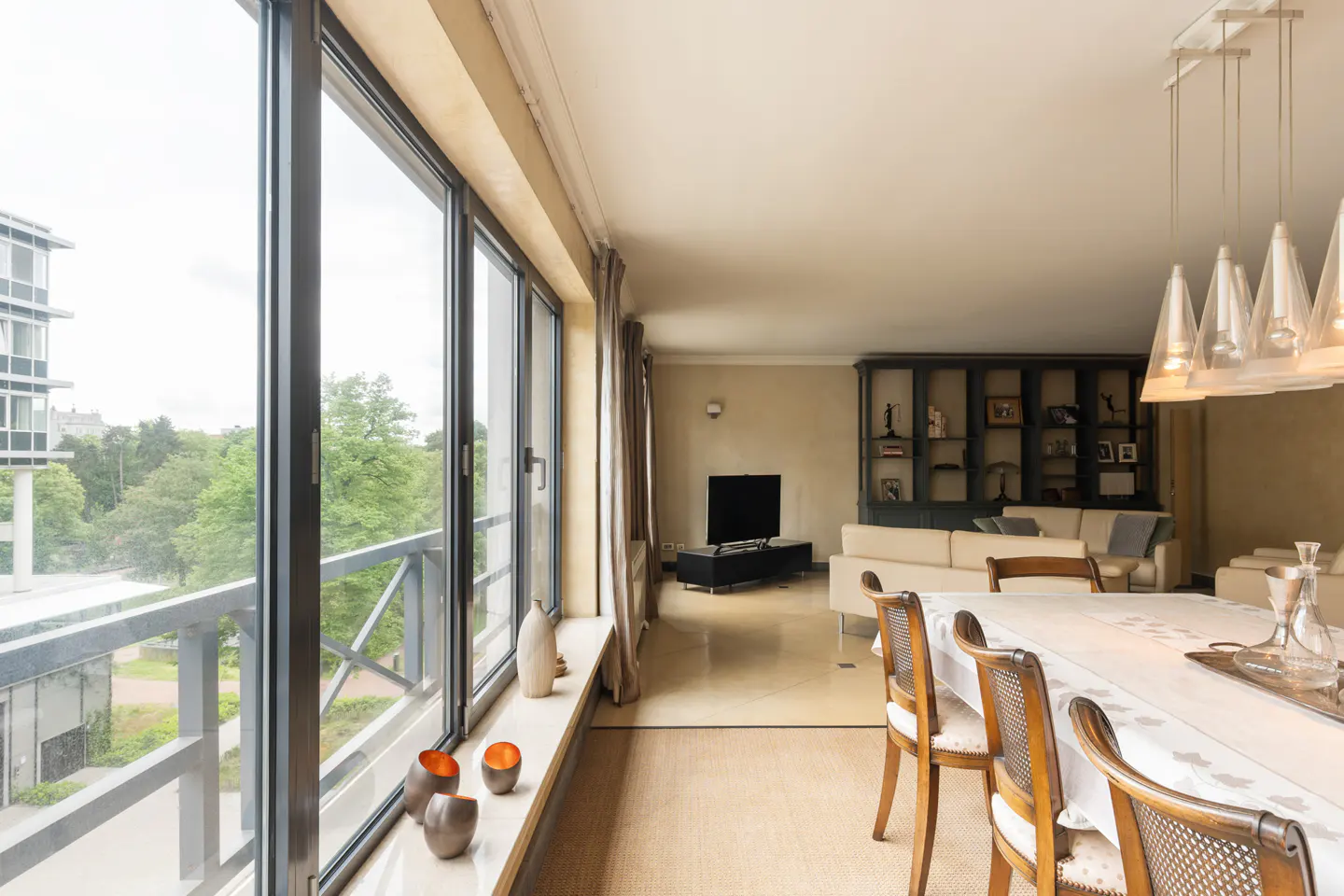Bright living room with large windows overlooking greenery. Dining table in foreground, sofa and TV in background.