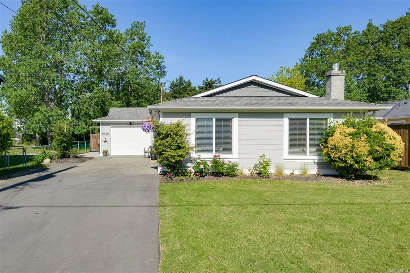 Single-story grey house with a white garage, paved driveway, and green lawn under a clear blue sky.