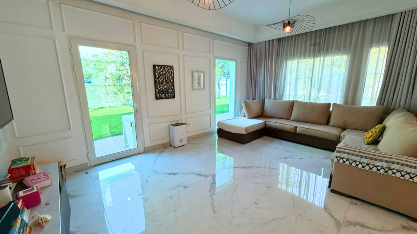 Bright living room with marble floors, a beige sectional sofa, and white paneled walls. Large windows offer a view of the green lawn outside.