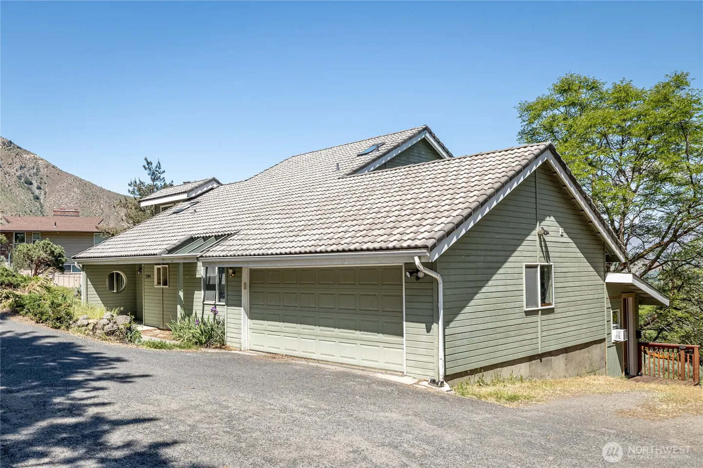 Exterior view of a green house with a gray tiled roof, a two-car garage, and a gravel driveway on a sunny day.