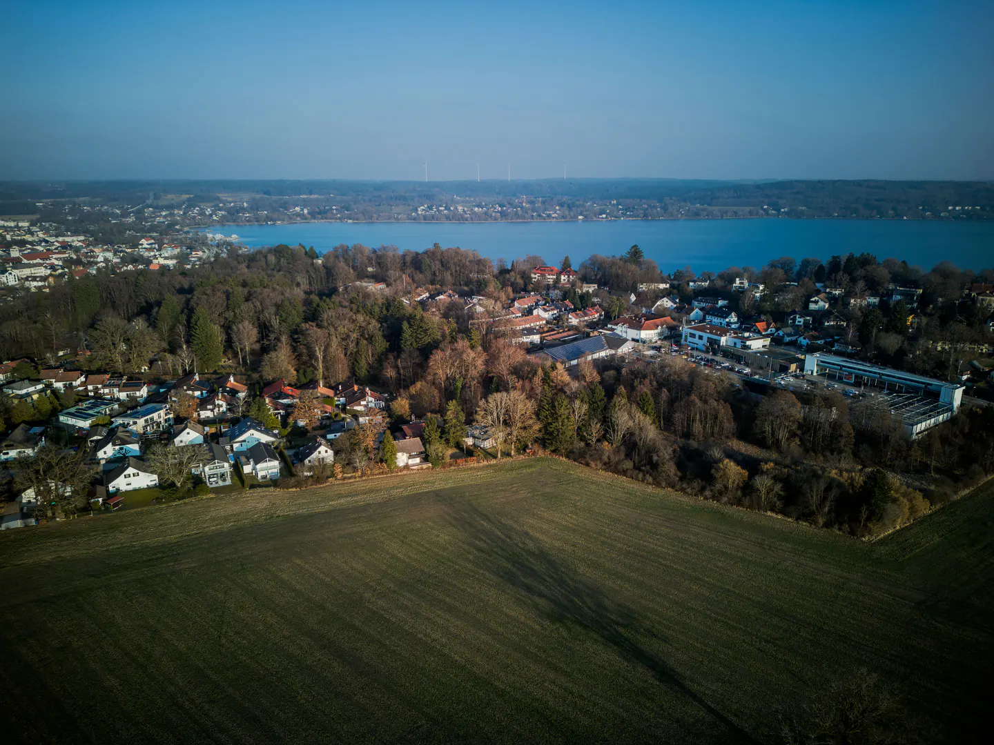 Aerial view of a town with houses, trees, and a green field in the foreground, with a blue lake and sky in the background.