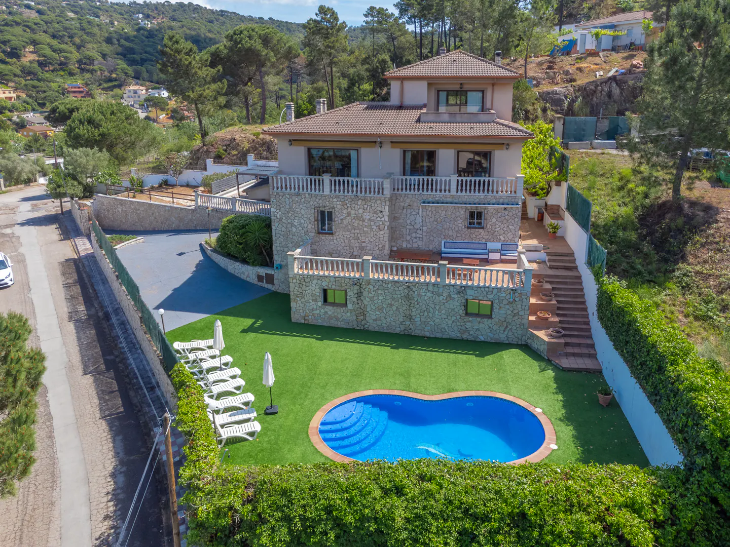 Aerial view of a two-story stone house with a pool, lawn chairs, and green trees in the background.