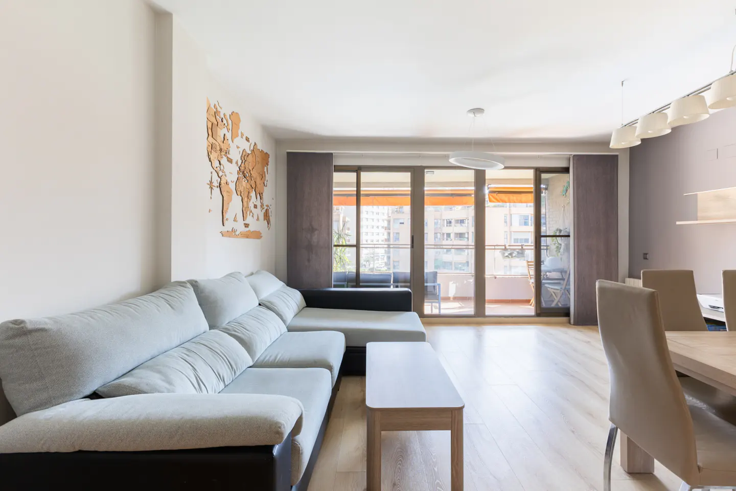 Bright living room with a gray sectional sofa, a wooden world map on the wall, and a balcony with city views.