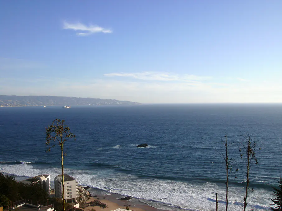 Ocean view with waves crashing on a sandy beach, buildings, and trees under a blue sky.