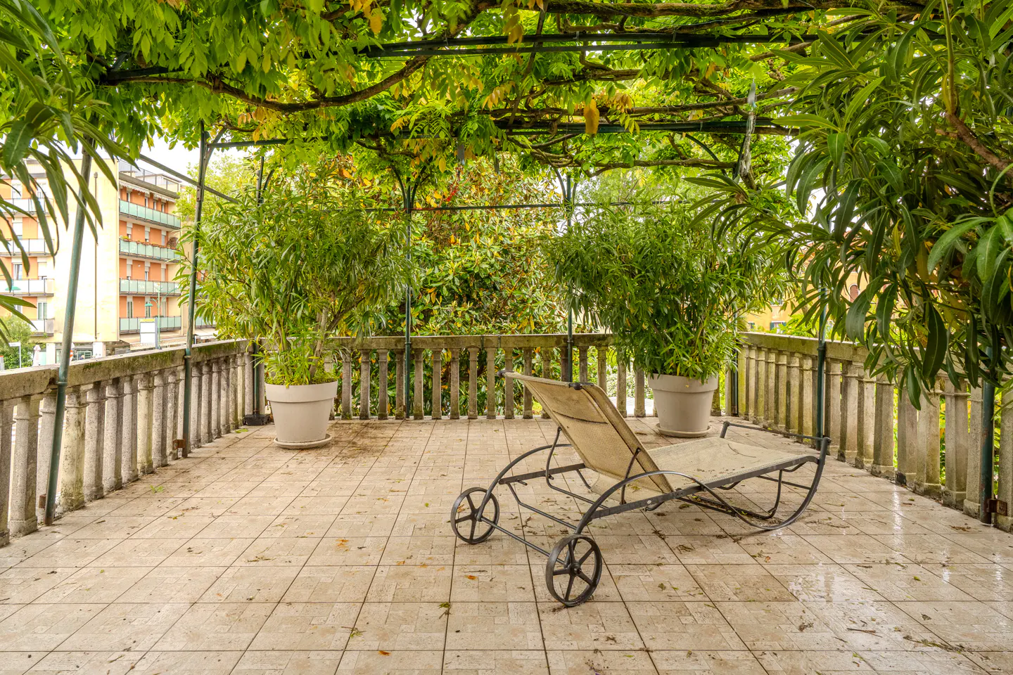 A sun lounger sits on a tiled patio with potted plants and a vine-covered pergola. A building is visible in the background.