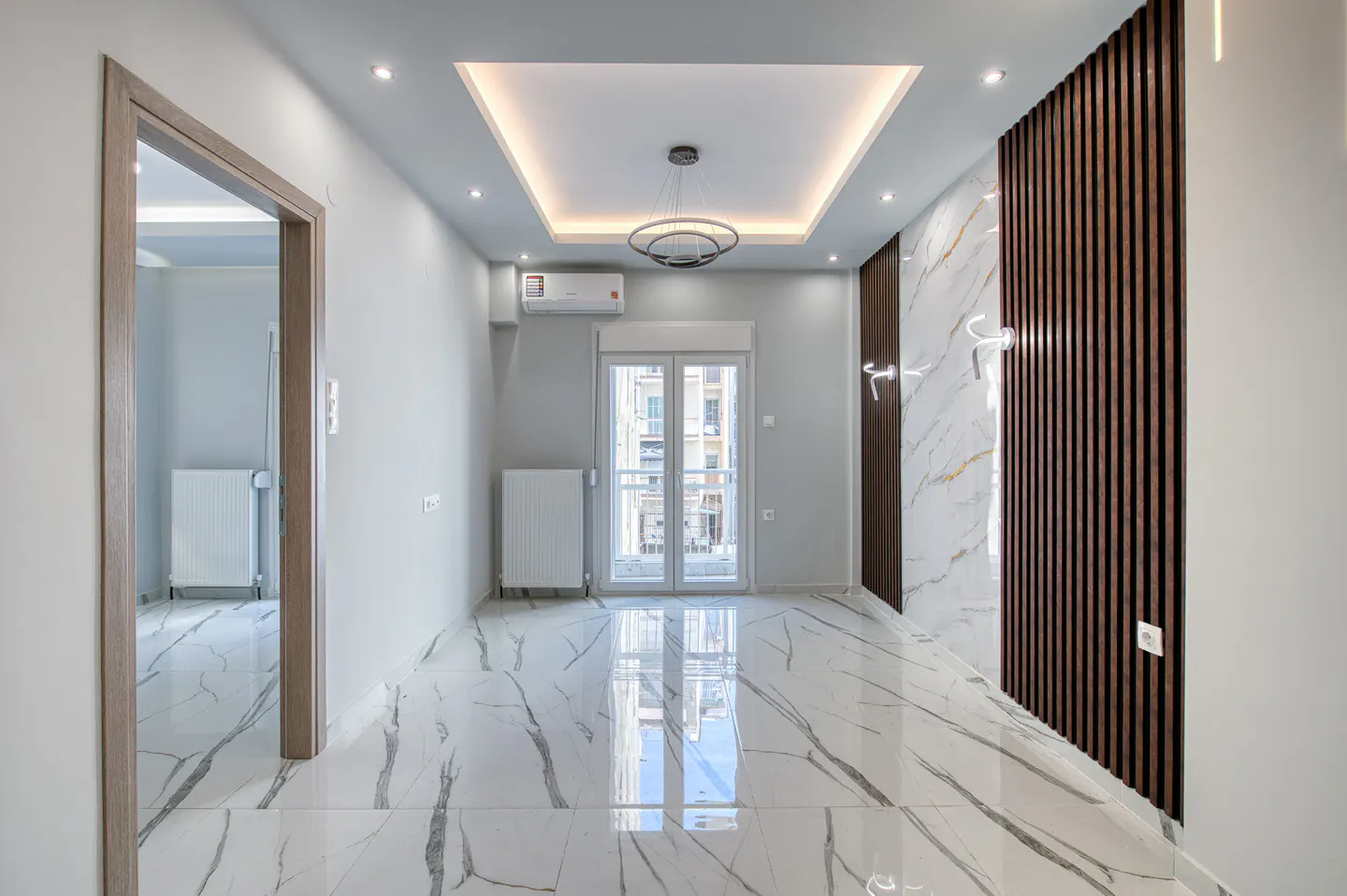 Bright, empty room with white marble floors, gray walls, and a modern chandelier. A door and window let in natural light.