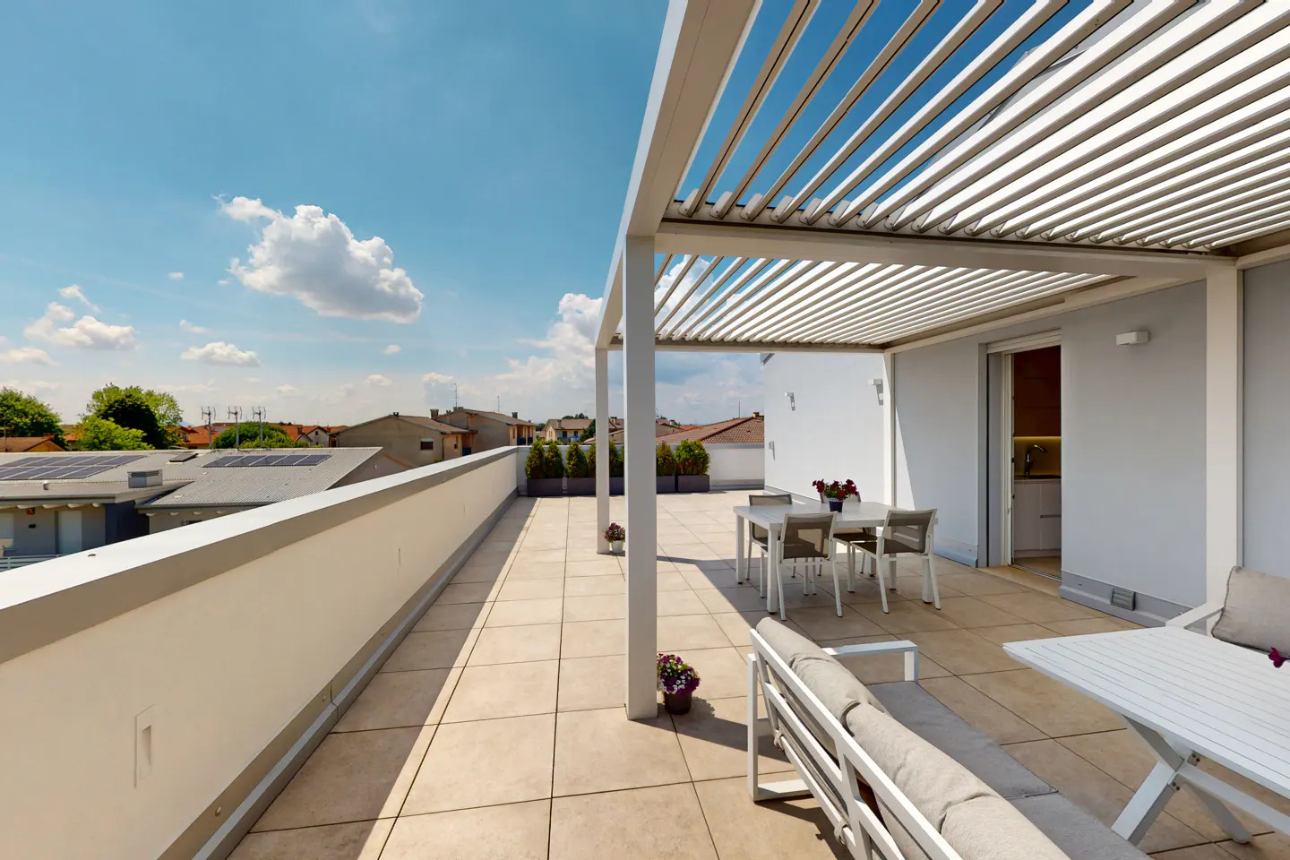 A rooftop patio with a white pergola, table, chairs, and a sofa. The patio has a low wall and overlooks a neighborhood with trees and blue sky.