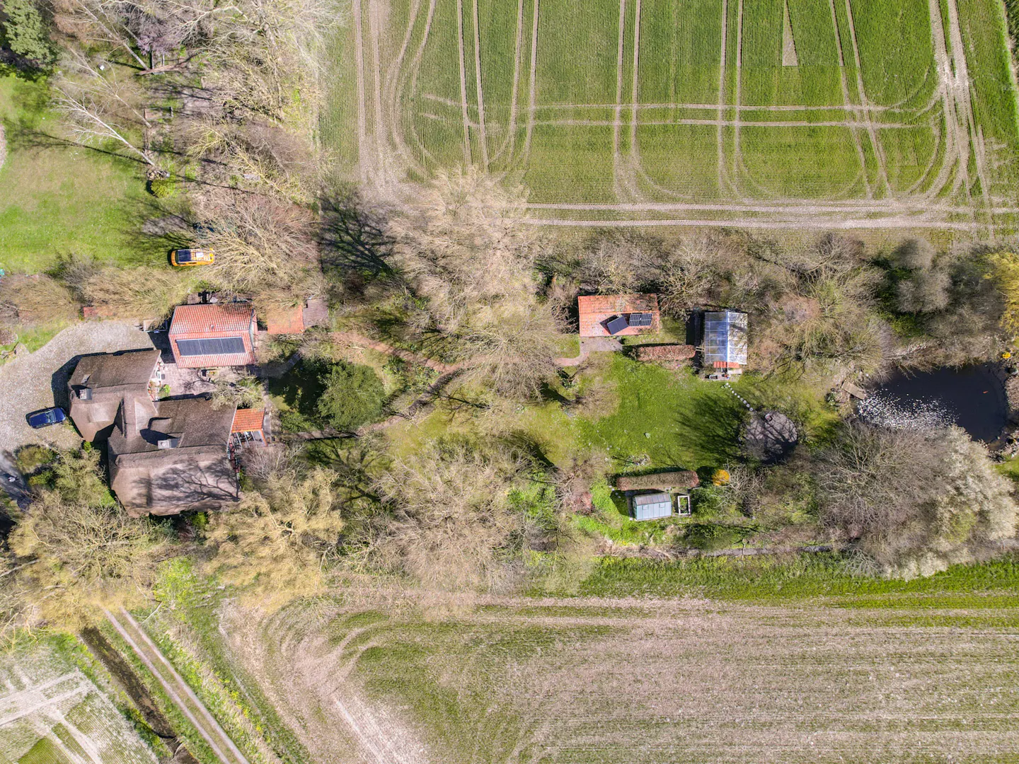 Aerial view of a rural property with a house, outbuildings, pond, and fields. The house has a dark roof and is surrounded by trees.