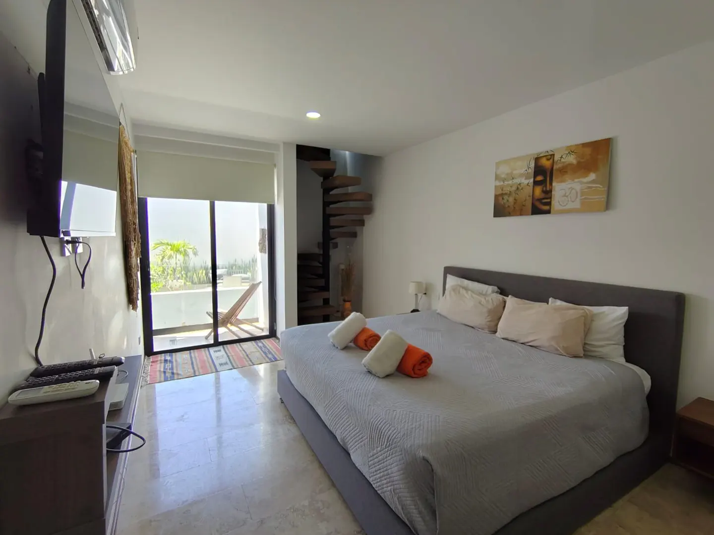 Bright bedroom with a gray bed, white and orange towels, and a spiral staircase. Balcony with a chair is visible through sliding glass doors.