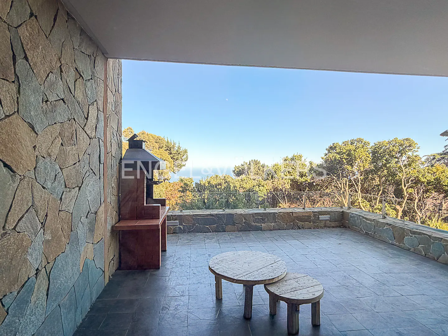 Outdoor patio with stone wall, grill, and two wooden tables overlooking trees and a blue sky.