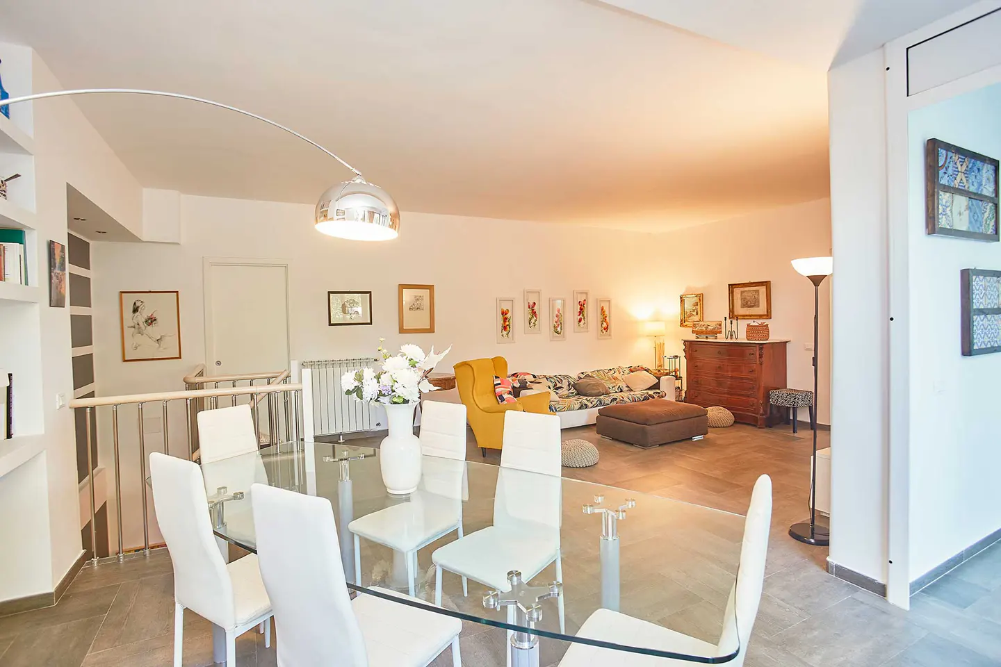 Bright open-plan living area with a glass dining table, white chairs, yellow armchair, and a brown sofa in the background.